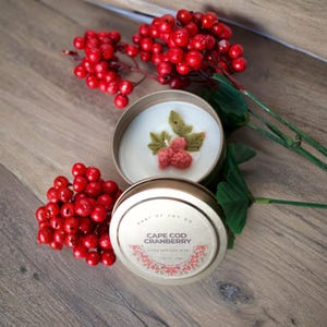May include: A gold-colored metal tin candle with a white wax surface featuring a decorative wax cranberry and leaf topper. The lid displays the text CAPE COD CRANBERRY, COCO APRICOT WAX, and 7 OZ (198G). Bright red artificial berry sprigs surround the candle.