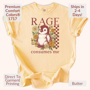 May include: Pale yellow t-shirt with a penguin graphic, the word "RAGE", and the phrase "consumes me". The design includes flowers and a checkered pattern. The shirt also has the text "Premium Comfort Colors 1717" and "Direct To Garment Printing".
