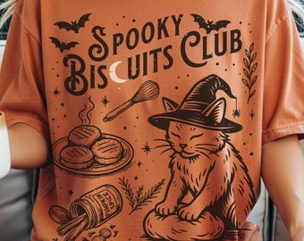 Op de afbeelding: Oranje T-shirt met een afbeelding van een kat met een heksenhoed, koekjes, vleermuizen en de tekst "Spooky Biscuits Club". Het ontwerp bevat een garde, kruiden en een halve maan. Het shirt is een comfortabele, casual stijl.