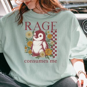 May include: A pale green t-shirt with a penguin illustration and the text "RAGE consumes me". The penguin is surrounded by yellow flowers, and the design includes a checkered pattern in a maroon colour.