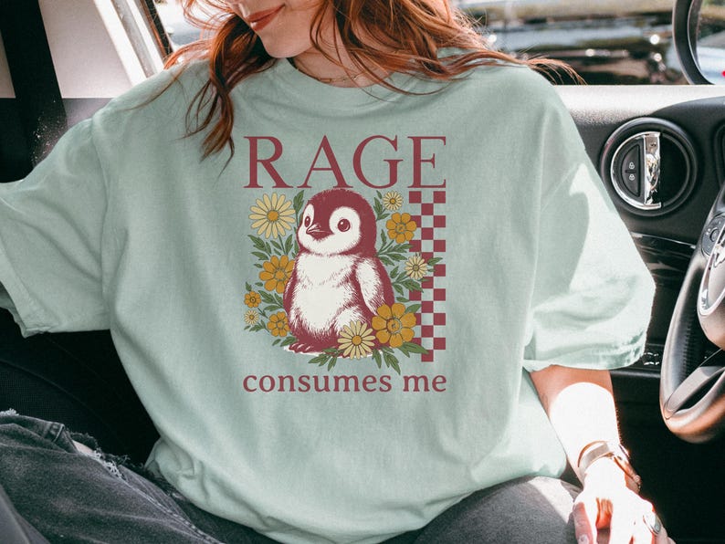 May include: A pale green t-shirt with a penguin illustration and the text "RAGE consumes me". The penguin is surrounded by yellow flowers, and the design includes a checkered pattern in a maroon colour.