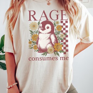 May include: A cream-coloured t-shirt with a penguin illustration surrounded by yellow and brown flowers. The text "RAGE consumes me" is printed above and below the penguin design. A checkered pattern is on the right side of the design.