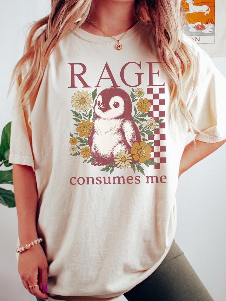 May include: A cream-coloured t-shirt with a penguin illustration surrounded by yellow and brown flowers. The text "RAGE consumes me" is printed above and below the penguin design. A checkered pattern is on the right side of the design.