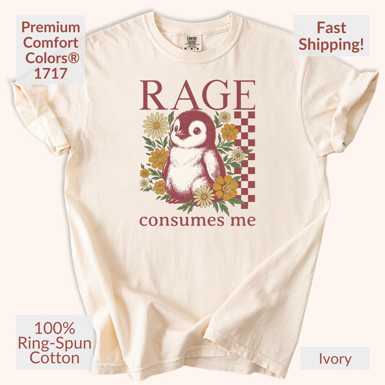 May include: Ivory t-shirt featuring a penguin graphic with the word "RAGE" and the phrase "consumes me". The design is surrounded by flowers. Made from 100% ring-spun cotton.