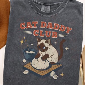 May include: Dark gray t-shirt with the text "CAT DADDY CLUB" in red and white. The shirt features a cartoon cat with blue eyes, kneading dough on a wooden board, with a bowl and eggs nearby. A fun, cat-themed graphic tee.