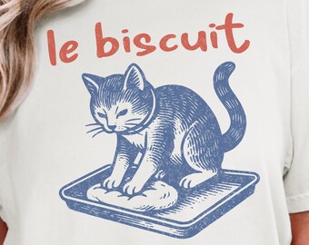 May include: Cream-colored t-shirt featuring a blue illustration of a cat kneading dough on a tray. The text "le biscuit" is printed in red above the cat. The design is a vintage-style graphic.
