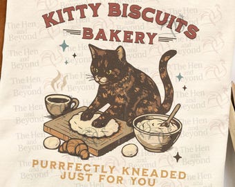 Puede incluir: Una camiseta color crema con un gráfico de un gato carey amasando masa sobre una tabla de madera. El texto dice KITTY BISCUITS BAKERY y PURRFECTLY KNEADED JUST FOR YOU. Incluye ilustraciones de una taza de café, un tazón y huevos.