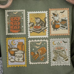 May include: Green sweatshirt featuring six postage stamp designs with Halloween themes. The stamps depict pumpkins, cats, baking, and ingredients like flour and pumpkin spice. Bats and spiderwebs add to the spooky aesthetic.