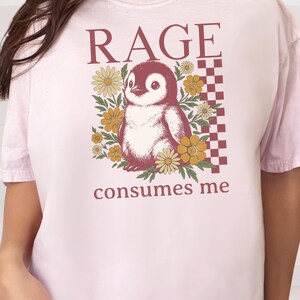 May include: A pale pink t-shirt with a penguin graphic surrounded by yellow flowers. The text "RAGE" is above the penguin, and "consumes me" is below. A checkered pattern is on the right side of the penguin.