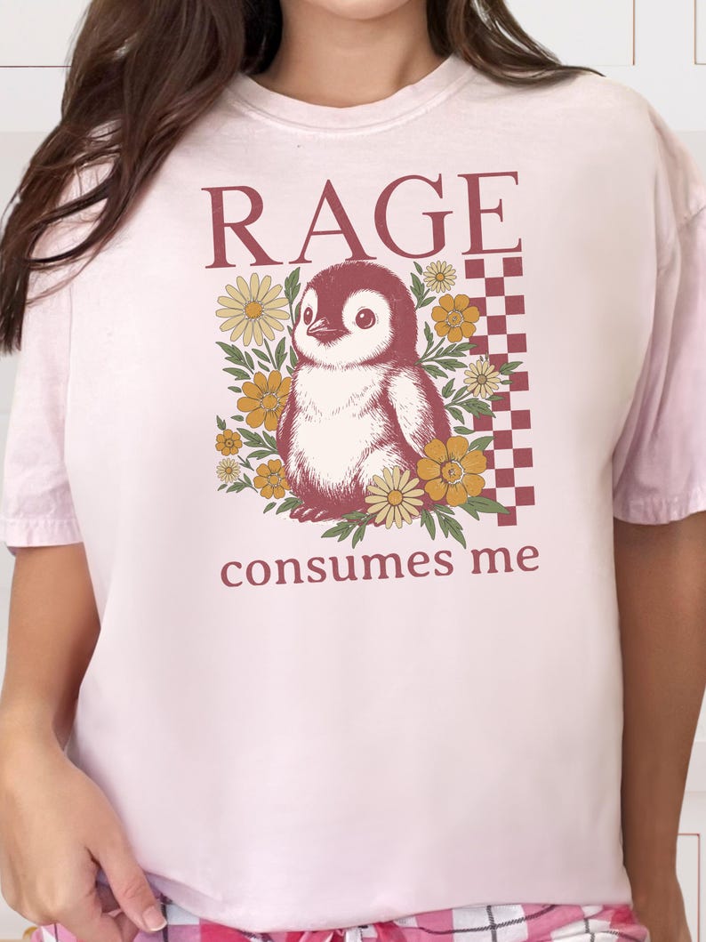 May include: A pale pink t-shirt with a penguin graphic surrounded by yellow flowers. The text "RAGE" is above the penguin, and "consumes me" is below. A checkered pattern is on the right side of the penguin.