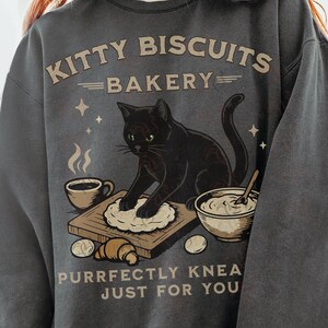 May include: Dark gray sweatshirt with a graphic of a black cat kneading dough on a wooden board. The text reads "Kitty Biscuits Bakery" above the cat and "Purrfectly Knea Just For You" below. A coffee cup, bowl, and eggs are also shown.
