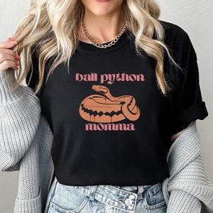 Op de afbeelding: Zwart T-shirt met een roze en oranje afbeelding van een ball python slang en de tekst "ball python momma".