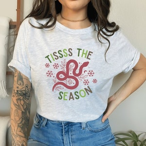 May include: A white t-shirt with a green and red graphic design featuring a snake with the text "Tiss the Season".