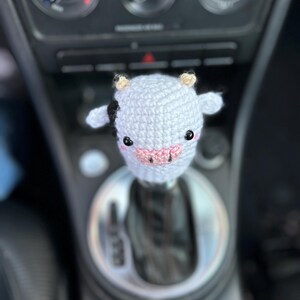 May include: A crocheted cow shift knob cover in light grey with black markings, small tan horns, and a pink nose. The cow has black eyes. The cover is designed to fit over a car's gear stick.