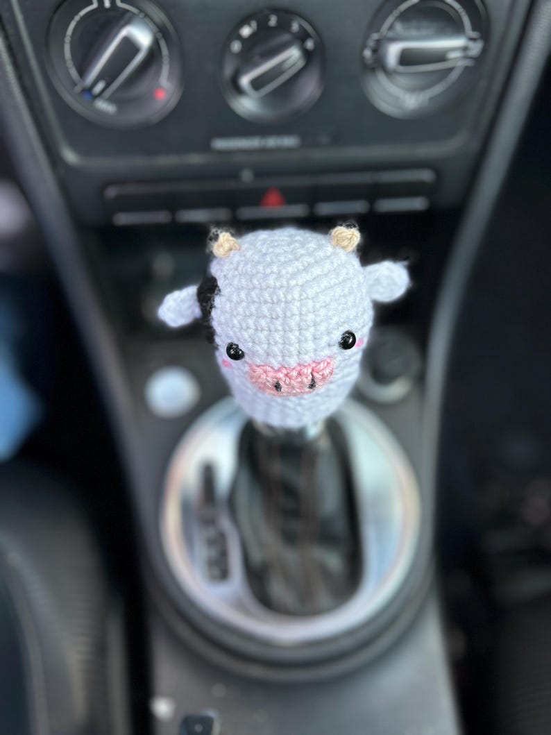 May include: A crocheted cow shift knob cover in light grey with black markings, small tan horns, and a pink nose. The cow has black eyes. The cover is designed to fit over a car's gear stick.