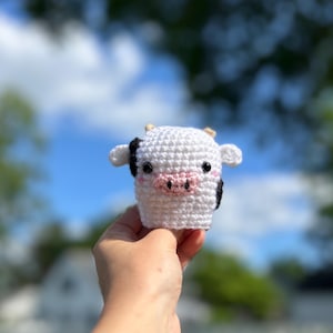 May include: A white crochet cow with black and pink details, including black ears and a pink snout. The handmade cow has small horns and is held in a hand against a blurred background of blue sky and green foliage.