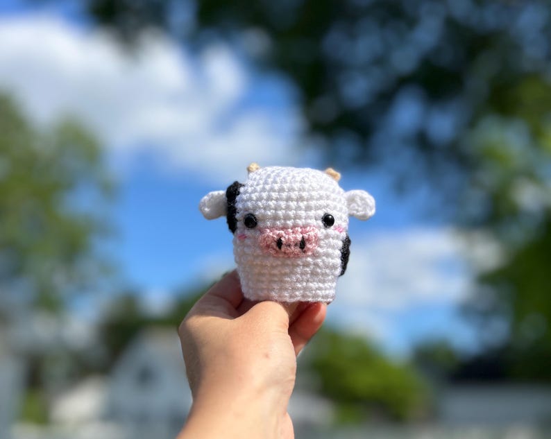 May include: A white crochet cow with black and pink details, including black ears and a pink snout. The handmade cow has small horns and is held in a hand against a blurred background of blue sky and green foliage.