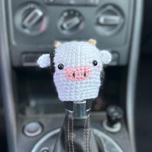 May include: A white and black crochet cow shift knob cover with a pink nose and black eyes. The handmade accessory is designed to fit over a car's gear shift lever. The cow has small ears and is attached to a black leather shift boot.