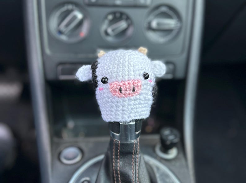 May include: A crochet cow shift knob cover in white and black, featuring a pink nose and black eyes. This handmade accessory is designed to fit over a car's gear shift lever. The cow has small ears and is attached to a black leather shift boot.