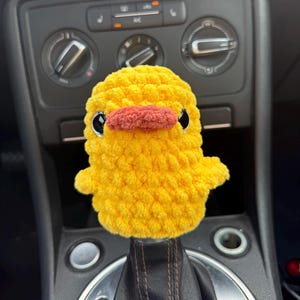 May include: A yellow crocheted duck cover for a gear shift knob, featuring a rounded body, small wings, and a dark orange beak. The duck has large, glossy black eyes with white highlights, positioned above the beak on the textured yarn surface.