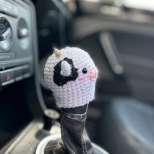 May include: A white and black crocheted cow shift knob cover with pink cheeks and small horns. The cover is placed over a black leather shift knob in a car.