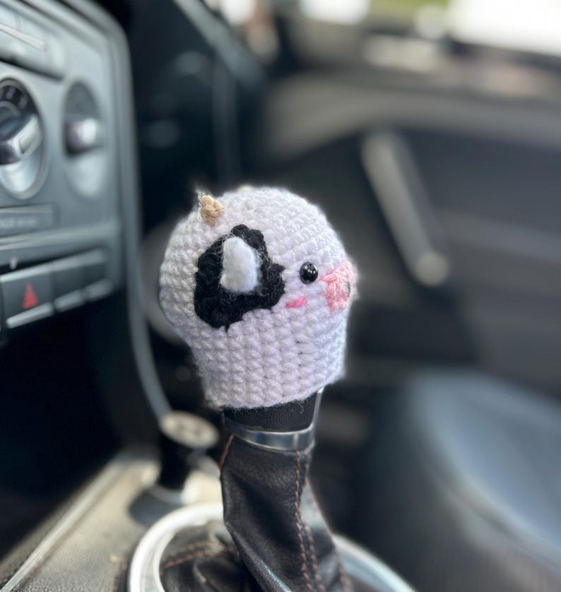 May include: A white and black crocheted cow shift knob cover with pink cheeks and small horns. The cover is placed over a black leather shift knob in a car.