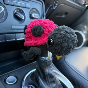 May include: A crochet ladybug gear shift cover featuring a bright red shell with black spots and a black head with shiny black plastic eyes. The plush, textured yarn accessory is fitted over the top of a black leather car gear shifter.