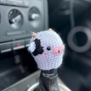 May include: A crocheted cow shift knob cover, white with black and pink details. The cover features a black patch, pink nose, and small horns. Designed to fit over a car's gear stick, it adds a decorative touch.