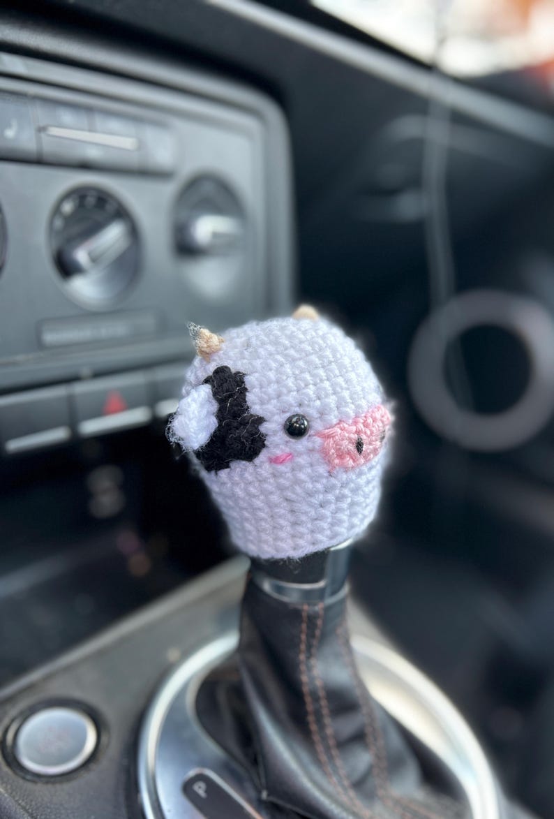May include: A crocheted cow shift knob cover, white with black and pink details. The cover features a black patch, pink nose, and small horns. Designed to fit over a car's gear stick, it adds a decorative touch.