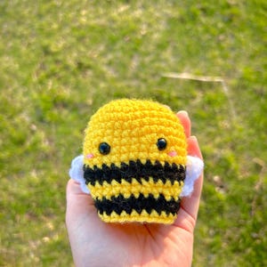 May include: A crocheted bee toy with black and yellow stripes, white wings, and black eyes. The bee is being held in a hand.
