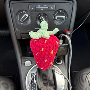May include: A crochet strawberry gear shift cover featuring a bright red body with small yellow embroidered seeds and a light green leafy top. The textured, yarn-based accessory is fitted over the gear shifter of a vehicle interior console.