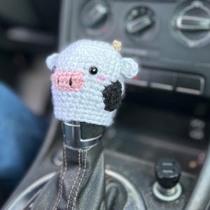 May include: A crocheted cow shift knob cover in white with black patches, a pink nose, and small, beige horns. This handmade cover is designed to fit over a car's gear stick, adding a touch of whimsy.