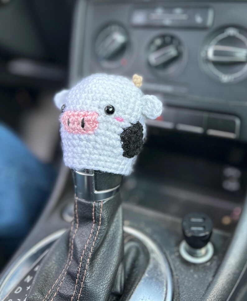 May include: A crocheted cow shift knob cover in white with black patches, a pink nose, and small, beige horns. This handmade cover is designed to fit over a car's gear stick, adding a touch of whimsy.