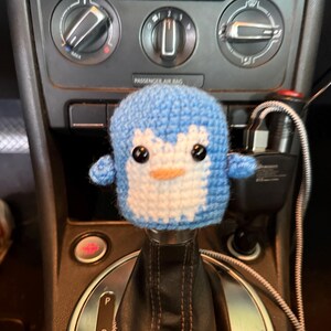 May include: A light blue and white crocheted penguin cover for a car gear shift knob. The penguin features two black plastic eyes, a small orange beak, and a rounded body shape that fits over the top of the gear shifter in a vehicle interior.
