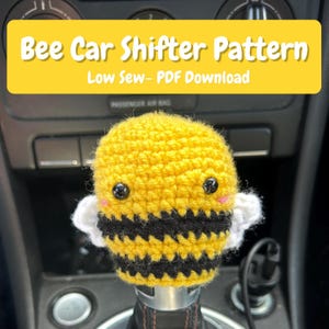 Könnte beinhalten: Ein gehäkelter Bienen-Schalthebelbezug mit gelben und schwarzen horizontalen Streifen, weißen Flügeln und schwarzen Sicherheitsaugen mit rosa Wangen. Der Text Bee Car Shifter Pattern Low Sew- PDF Download ist darüber zu sehen.