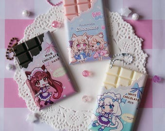 Könnte beinhalten: Drei rechteckige Schokoladentafel-Schlüsselanhänger mit Anime-Charakter-Illustrationen auf den Verpackungen. Eine ist rosa mit zwei Charakteren mit Fuchsohren, eine dunkelbraun mit einem Charakter im Dienstmädchen-Outfit und eine weiß.