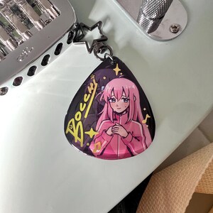 May include: A black guitar pick-shaped keychain featuring an anime character in a pink jacket. The keychain has a star-shaped clasp and the word "Bocchi" in gold lettering. The background is a light blue guitar.