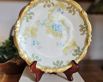 Pode incluir: Um prato decorativo antigo com uma borda dourada ondulada e design floral. O prato apresenta um fundo creme com detalhes florais em azul claro e amarelo. É exibido em um suporte de madeira.