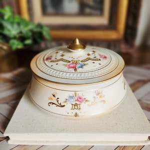May include: A cream-colored, lidded ceramic dish with floral and gold accents. The lid features pink and blue roses and a gold finial. The dish is likely vintage or antique, and is sitting on a cream-colored surface.