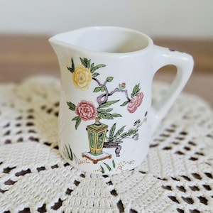 Könnte beinhalten: Ein weißes Milchkännchen aus Keramik mit einem bunten Blumen- und Laternenmuster. Das Design umfasst gelbe und rosa Blüten an grünen Stielen, eine grüne und gelbe Laterne auf einem kleinen Ständer sowie einen geschwungenen Henkel an der rechten Seite.