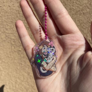 May include: A clear keychain with a holographic finish featuring a cartoon illustration of a person with long brown hair holding a cat. The person is wearing a dark blue shirt with a green star on it. The keychain has a pink chain.