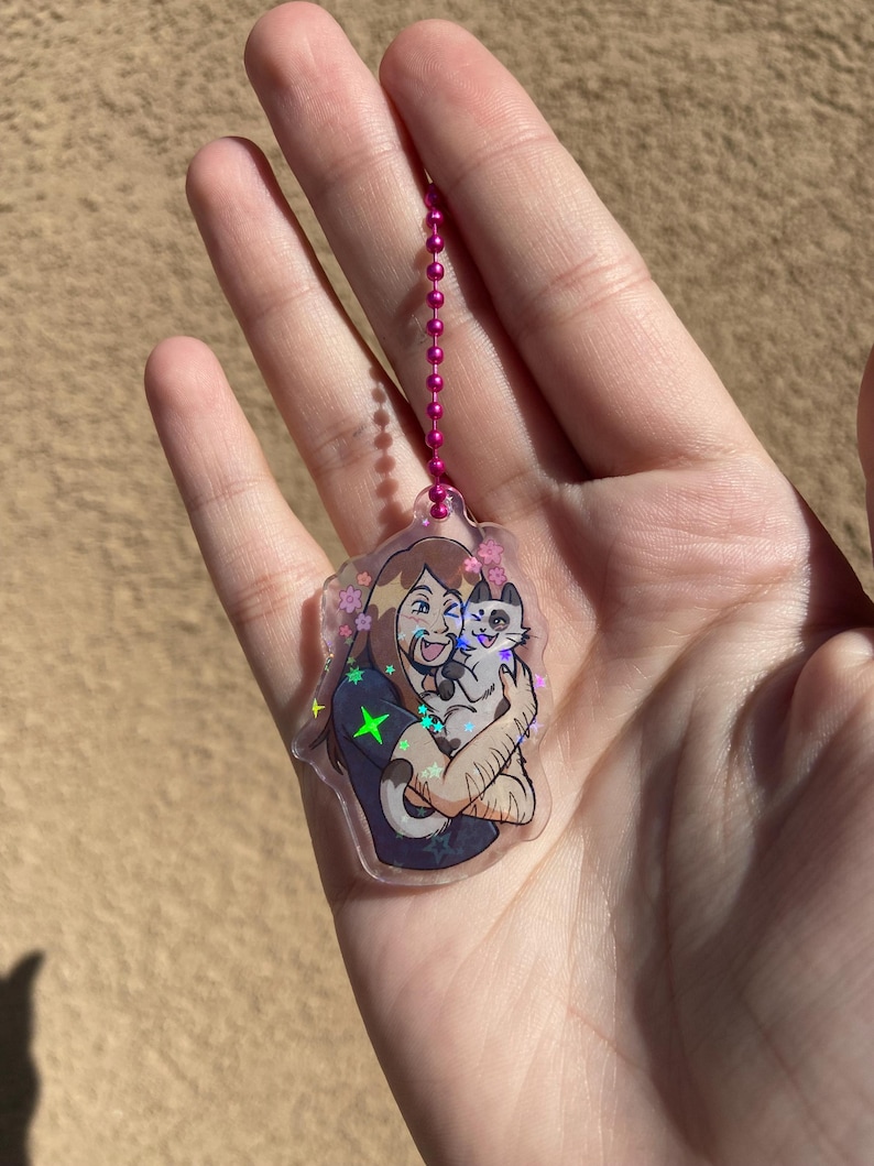 May include: A clear keychain with a holographic finish featuring a cartoon illustration of a person with long brown hair holding a cat. The person is wearing a dark blue shirt with a green star on it. The keychain has a pink chain.