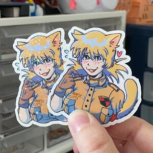 Puede incluir: Dos pegatinas de un gato de dibujos animados que lleva un uniforme amarillo y azul con el texto "Nurse Kitty" en el cuello. El gato tiene un rubor rosa en las mejillas y saca la lengua. Las pegatinas tienen un acabado hologr&aacute;fico.