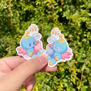 May include: Two stickers featuring a blue cartoon creature with wings, holding a pink laptop. The creature has a yellow antenna and a star-shaped sparkle. Text bubbles above the creature read "(232)" and "(2us)". The stickers are held against a green, leafy background.