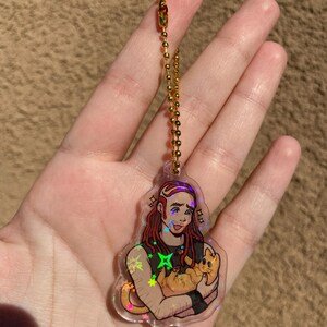May include: A holographic keychain featuring a cartoon illustration of a person with long red hair holding a cat. The keychain has a gold chain.