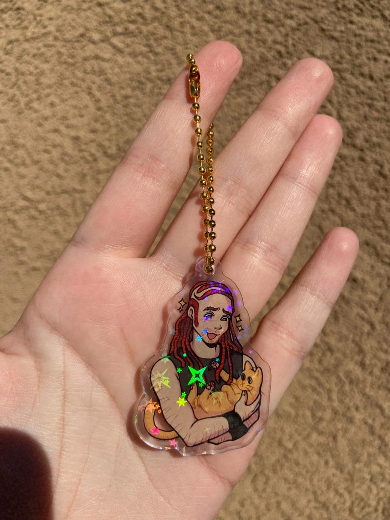 May include: A holographic keychain featuring a cartoon illustration of a person with long red hair holding a cat. The keychain has a gold chain.