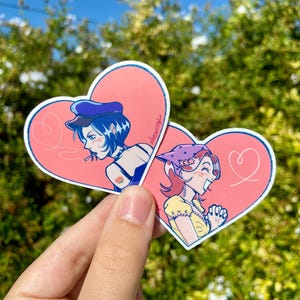 May include: Two heart-shaped stickers featuring illustrations of women. One woman wears a blue hat and smokes a cigarette, the other wears a headscarf and a yellow dress. Both are set against a pink background.
