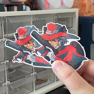 Puede incluir: Dos pegatinas hologr&aacute;ficas con un personaje de dibujos animados con cabello rojo, un sombrero rojo y una capa roja. El personaje sostiene una pistola y tiene un pentagrama en el pecho. El texto "Muse Kitty" es visible en la pegatina.