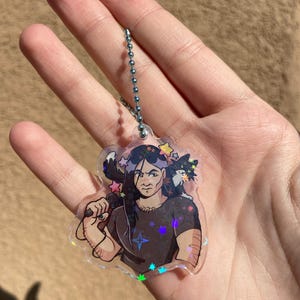 May include: A holographic keychain featuring a cartoon illustration of a person with long black hair holding a microphone. The person is surrounded by stars and a black cat with white markings.