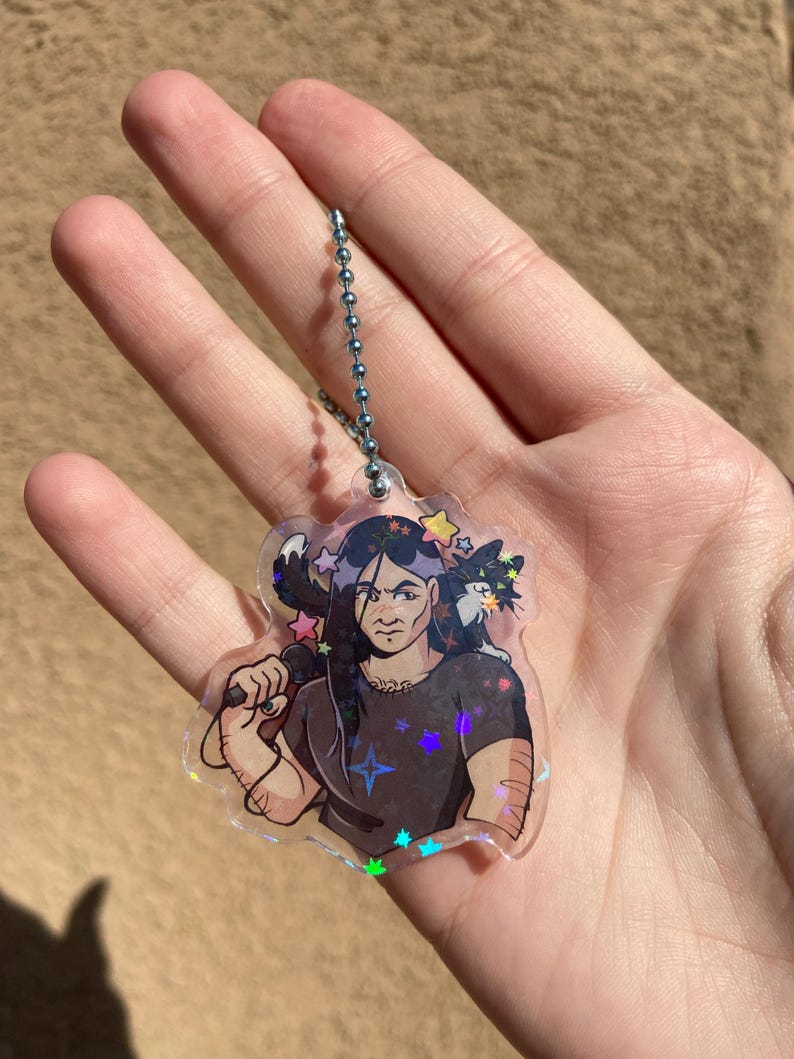 May include: A holographic keychain featuring a cartoon illustration of a person with long black hair holding a microphone. The person is surrounded by stars and a black cat with white markings.
